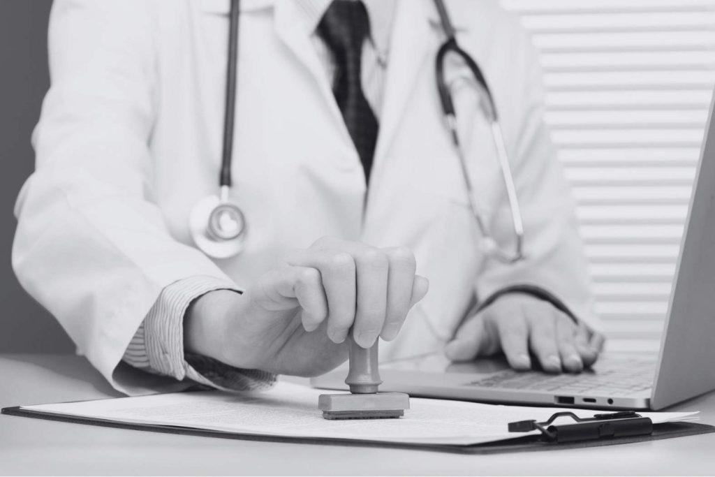 Médico estampando un sello en documento de homologación médica frente a un computador portátil