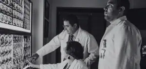 Tres médicos con bata blanca analizan imágenes radiológicas en un negatoscopio dentro de un hospital, en blanco y negro