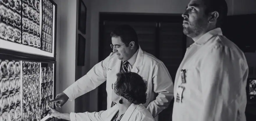 Tres médicos con bata blanca analizan imágenes radiológicas en un negatoscopio dentro de un hospital, en blanco y negro