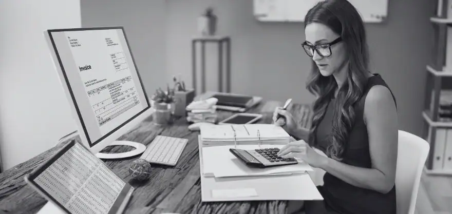 Mujer con gafas usa una calculadora frente a documentos y un monitor con una factura en un escritorio de oficina, en blanco y negro