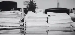 Pilas de documentos y carpetas apiladas sobre un escritorio de oficina en blanco y negro, con sillas al fondo