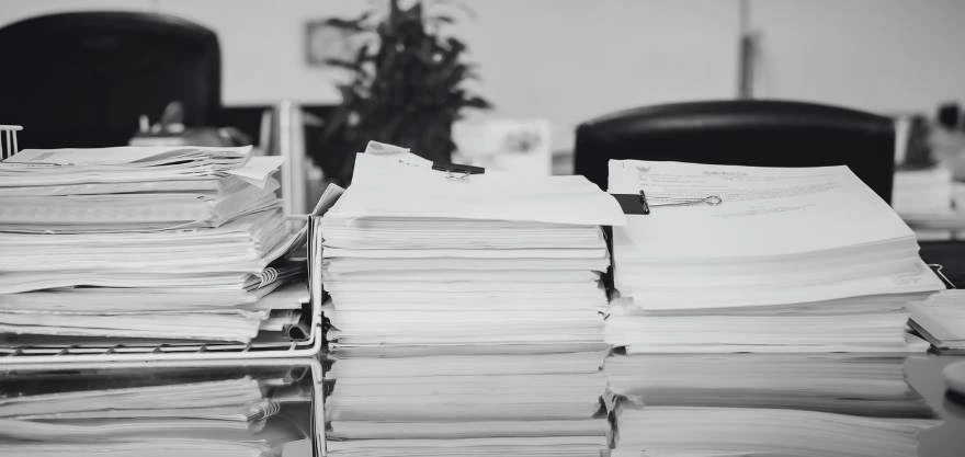 Pilas de documentos y carpetas apiladas sobre un escritorio de oficina en blanco y negro, con sillas al fondo