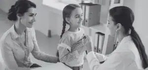 Médica con estetoscopio atiende a una niña sonriente acompañada de una mujer en una consulta, en fotografía en blanco y negro