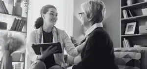 Médica con bata y tableta conversa sonriente con una mujer mayor sentada en un sofá, en fotografía en blanco y negro